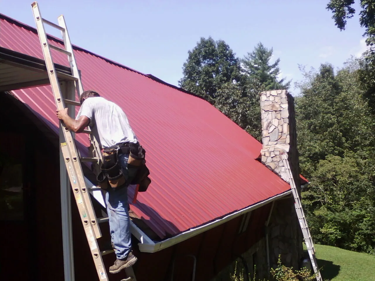 Roofer on a ladder beside a red metal roof and stone chimney — Soffit & Fascia Repair by Roofing Brothers in Ceresco